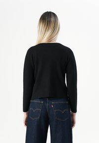 Une personne avec de longs cheveux blonds porte un haut noir à manches longues et un jean en denim bleu foncé, de dos sur un fond blanc.