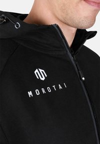 Čierna zipsová bunda vyrobená z mäkkého materiálu. Má vysoký golier, nastaviteľnú kapucňu a biele logo "MOROTAI" na hrudi.