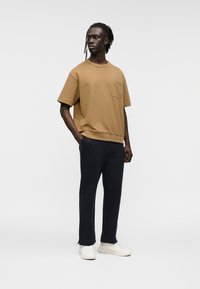 Καστανό βαμβακερό μπλουζάκι με τσέπη, συνδυασμένο με ναυτικού μπλε παντελόνια και λευκά αθλητικά παπούτσια. Το μπλουζάκι έχει άνετη εφαρμογή και στρογγυλή λαιμόκοψη.