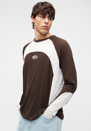 Brown and white long-sleeve T-shirt with a ribbed texture, featuring contrasting panels and a logo on the chest. Relaxed fit.