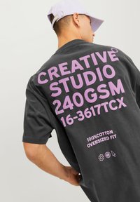 Tamno siva oversized majica s podebljanim ružičastim tekstom na leđima, s natpisom "CREATIVE STUDIO 240GSM 16-3617TCX" i detaljima o tkanini.