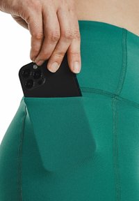 Leggings deportivos verdes con un bolsillo lateral que sostiene un teléfono inteligente negro, mostrando un diseño elegante y una textura suave.