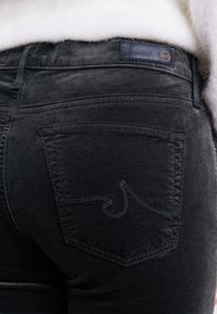 Černé denimové kalhoty mají zadní kapsu s zaobleným švem. Tkanina má texturovaný vzhled a na pase je viditelná kožená nášivka.