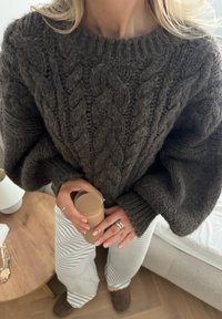 Un pull en tricot gris foncé, épais, avec un motif en cable et des manches surdimensionnées. Pantalons rayés blancs, tenant un verre transparent avec une boisson légère.