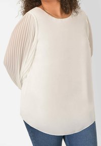 Light beige top with pleated sheer sleeves, rounded neckline, and a flowing design. Fabric appears smooth with a soft drape, paired with denim.