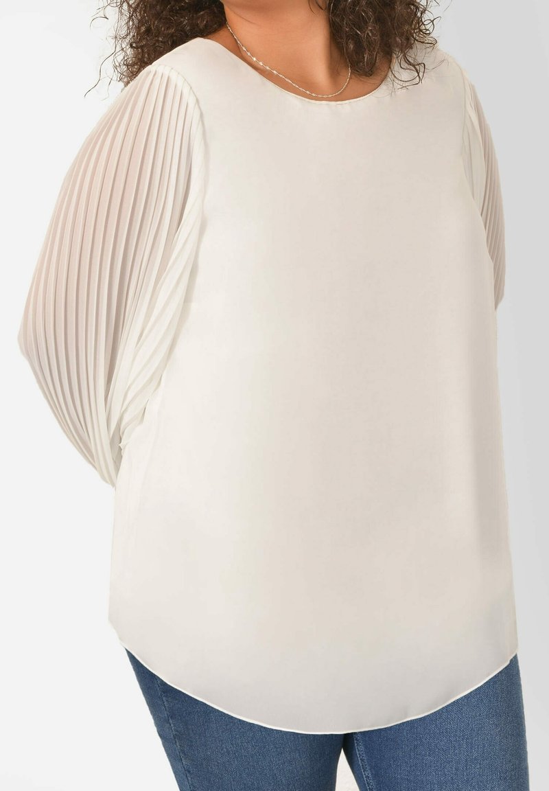 Light beige top with pleated sheer sleeves, rounded neckline, and a flowing design. Fabric appears smooth with a soft drape, paired with denim.