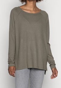 Person in einem olivgrünen Langarmpullover und hellgrauen Jeans, der vor einem einfarbigen hellen Hintergrund steht.