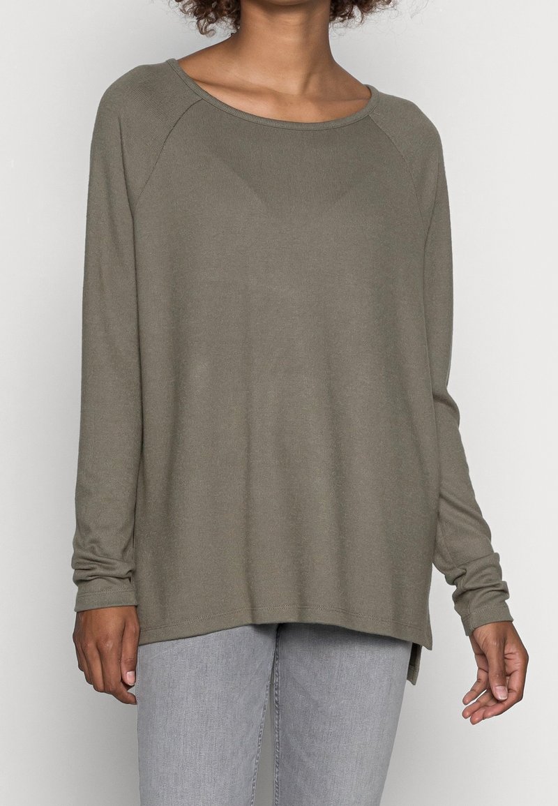 Person in einem olivgrünen Langarmpullover und hellgrauen Jeans, der vor einem einfarbigen hellen Hintergrund steht.
