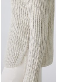 Hellbeige gestrickter Pullover mit Rippstruktur, langen Ärmeln und seitlichen Schlitzen, vor einem neutralen Hintergrund gezeigt.