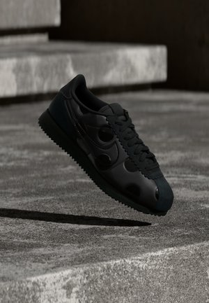 Zwarte sneaker met een geribbelde zool en een cirkelvormig detail aan de zijkant, hangend boven een betonnen oppervlak met trappen op de achtergrond.