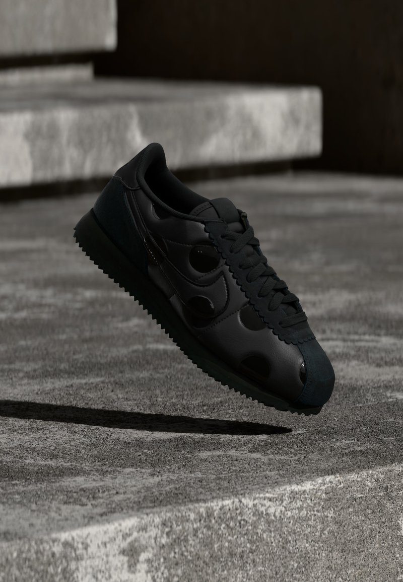 Μαύρο αθλητικό παπούτσι με ραβδωτή σόλα και κυκλικό διακοσμητικό στη πλευρά, αιωρούμενο πάνω από μια επιφάνεια από σκυρόδεμα με σκάλες στο φόντο.