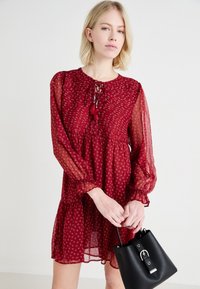 Rochie roșie cu model floral, cu mâneci lungi din material transparent, decolteu cu fundă și fustă în straturi. Acoperită de o geantă neagră de tip bucket cu accesorii din argint.