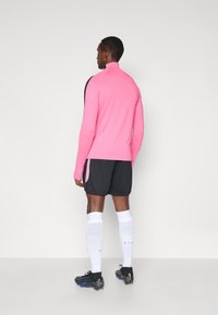 Ροζ αθλητικό μπλουζάκι με μακριά μανίκια, μαύρο σορτς, λευκές κάλτσες μέχρι το γόνατο και μαύρα ποδοσφαιρικά παπούτσια. Λείο ύφασμα, εφαρμοστή σχεδίαση και αντίθετα χρώματα.