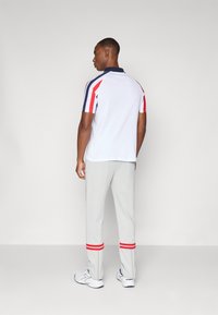 Weißes Poloshirt mit marineblauem Kragen und rot-blauen Schulterstreifen, kombiniert mit grauen Trainingshosen, die rote Akzente an den Bündchen aufweisen.