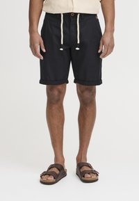 Pantaloni corti neri in lino con risvolti, tasche frontali e un cordoncino beige a contrasto, indossati con sandali in pelle marrone.