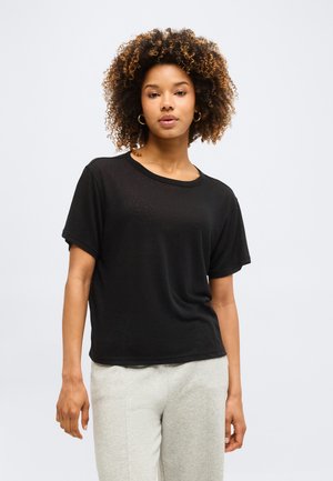 Γυναίκα με σγουρά μαλλιά που φοράει χρυσούς κρίκους στα αυτιά, μαύρο κοντομάνικο πουκάμισο και ανοιχτό γκρι παντελόνια σε ένα απλό φόντο.