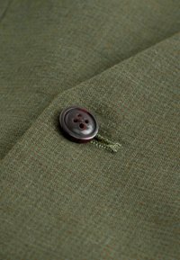 Bouton marron foncé cousu sur un tissu texturé vert-brun avec des coutures visibles et une boutonnière à côté.