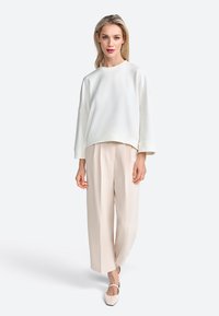 Felpa bianca, oversize, con spalle basse abbinata a pantaloni beige chiaro a pieghe e scarpe con punta, su uno sfondo semplice.