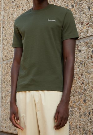 Homme portant un t-shirt à manches courtes Calvin Klein vert olive et un pantalon ample beige clair, debout contre un mur en pierre texturé.