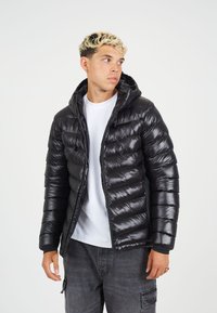 Črna napihnjena jakna z bleščečo površino, vodoravno prešita zasnova, sprednji zadrga in pritrjena kapuca, oblečena čez belo majico.