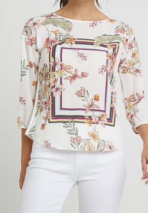 Biała bluzka z kolorowymi kwiatowymi wzorami i kwadratowym paskowanym obramowaniem, zestawiona z białymi spodniami, pokazana od ramion do bioder.