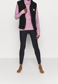 Musta fleece-takki vaaleanpunaisen pitkähihasen paidan päällä, jossa on tähden kuvio, yhdistettynä mustiin legginsseihin. Paljaat jalat näkyvissä. Paidassa on sivutasku.