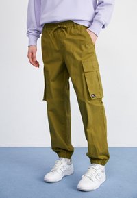 Olive zielone spodnie bojówki z elastycznym pasem, dwoma bocznymi kieszeniami i elastycznymi mankietami. Noszone z białymi tenisówkami, prezentują casualowy wygląd.