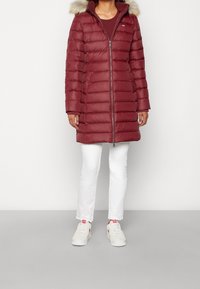 Abrigo rojo tipo puffer con cierre frontal de cremallera y capucha con borde de piel. Presenta secciones acolchadas horizontales y una silueta ajustada, combinado con pantalones blancos y zapatillas.