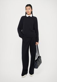 Maglione a maglia blu navy con colletto bianco, abbinato a pantaloni neri a righe sottili. La modella tiene una borsa nera e indossa scarpe bianche con tacco a blocco.