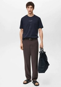 Navy cotton t-shirt with printed text, brown linen trousers, black belt, black bag with straps, and black slide sandals.