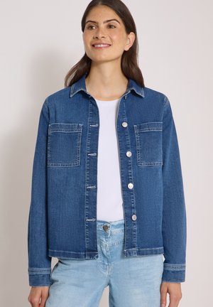 Femme portant une veste en jean bleu foncé sur une chemise blanche et un jean bleu clair, souriante et regardant légèrement vers la droite.