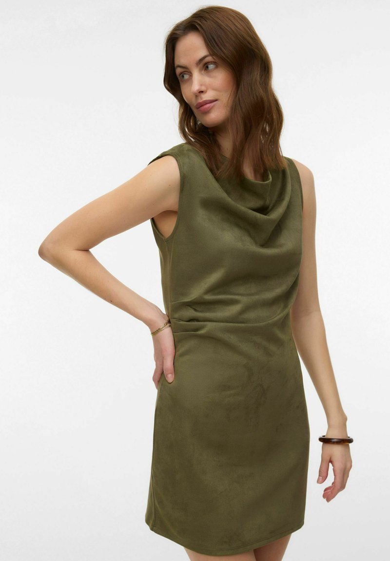 Femme aux cheveux bruns mi-longs portant une robe sans manches vert olive avec un décolleté drapé, debout avec une main sur la hanche.