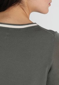 Nahaufnahme des oberen Rückens und Nackens einer Frau, die einen dunkelgrauen Pullover mit einem weißen Streifenkragen trägt.