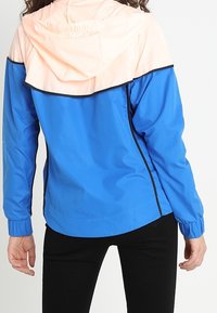 Blå lättviktsjacka med en persiko-färgad panel upptill, utrustad med huva, elastiska ärmslut och svart kant längs sömmarna.
