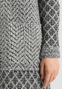 Suéter de punto en blanco y negro, con un patrón texturizado y puños grises texturizados. El diseño es intrincado y de múltiples capas.