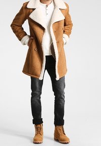 Brun sherlingskappa med en vit pälskrage och manschetter, parat med mörka jeans och ljusbruna snörstövlar. Kappan har en dubbelknäppt design.