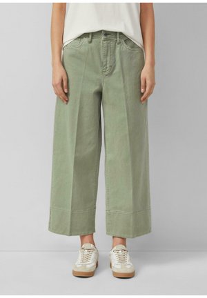 Femme portant un pantalon ample mi-long vert olive clair et des baskets blanches, debout devant un fond uni gris clair.