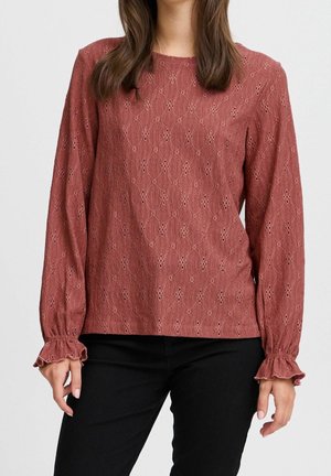 Long-sleeve rust blouse with small eyelet details, round neckline, and ruffled cuffs worn with black pants.