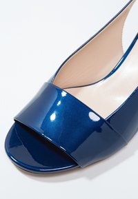 Chaussure ouverte bleu marine brillante avec large bride avant et doublure intérieure beige sur fond blanc.