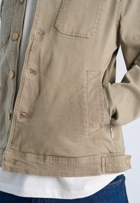 Close-up of beige casual jacket with front buttons, chest and side pockets, worn over white shirt and paired with blue jeans.