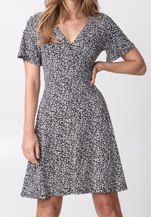 Femme en robe noire à col en V avec imprimé floral blanc petit, manches courtes, et jupe évasée, debout avec une main sur la hanche.