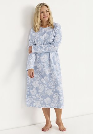 Kvinna står barfota mot en vit vägg iförd en blå långärmad klänning med vitt blommönster, ler och tittar åt vänster.