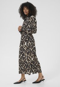 Abito maxi in tessuto nero con un motivo astratto beige e marrone a vortice. Presenta maniche lunghe, un colletto e uno spacco laterale. Abbinato a tacchi a molla neri.