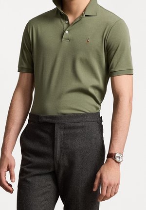 Man wearing olive green short-sleeve polo shirt and dark gray tailored pants, with a brown leather-strap wristwatch.