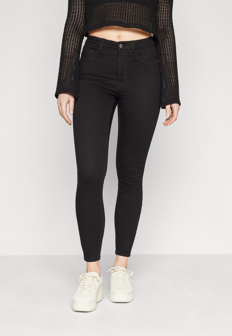 Mujer vestida con jeans ajustados negros de cintura alta, top de malla negro de manga larga y zapatillas de plataforma blancas, contra un fondo plano.