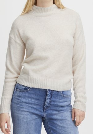 Femme portant un pull en maille couleur crème avec col et poignets côtelés, associé à un jean bleu en denim.
