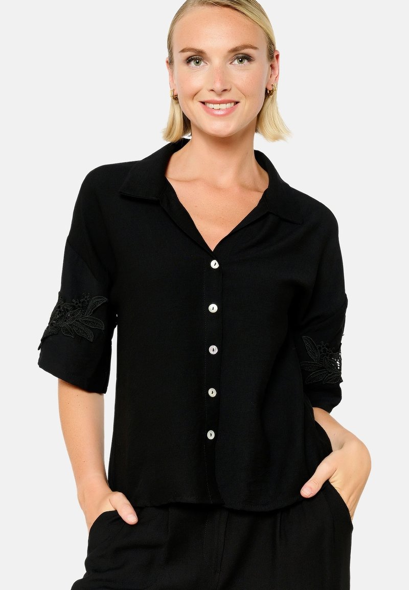 Blusa nera con bottoni e maniche corte, caratterizzata da un intricato ricamo nero sui polsini. Tessuto morbido, vestibilità comoda e design del colletto classico.