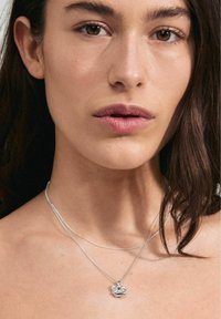 Collana d'argento con un pendente testurizzato, caratterizzato da due catene sottili e un centro trasparente e sfaccettato, su pelle nuda.