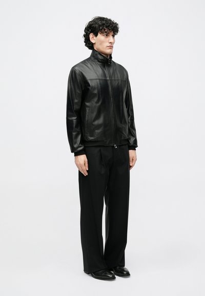 Crna kožna jakna s visokim ovratnikom, prednjim patentnim zatvaračem, dva bočna džepa i elegantnim završnim slojem, uparena s širokim crnim hlačama i cipelama.