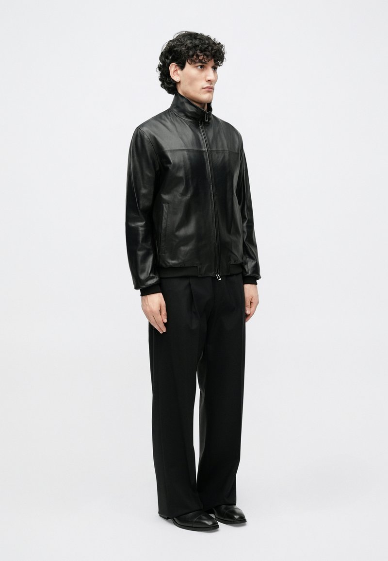 Veste en cuir noire à col montant, fermeture éclair frontale, deux poches latérales, finition élégante, associée à un pantalon noir ample et des chaussures.